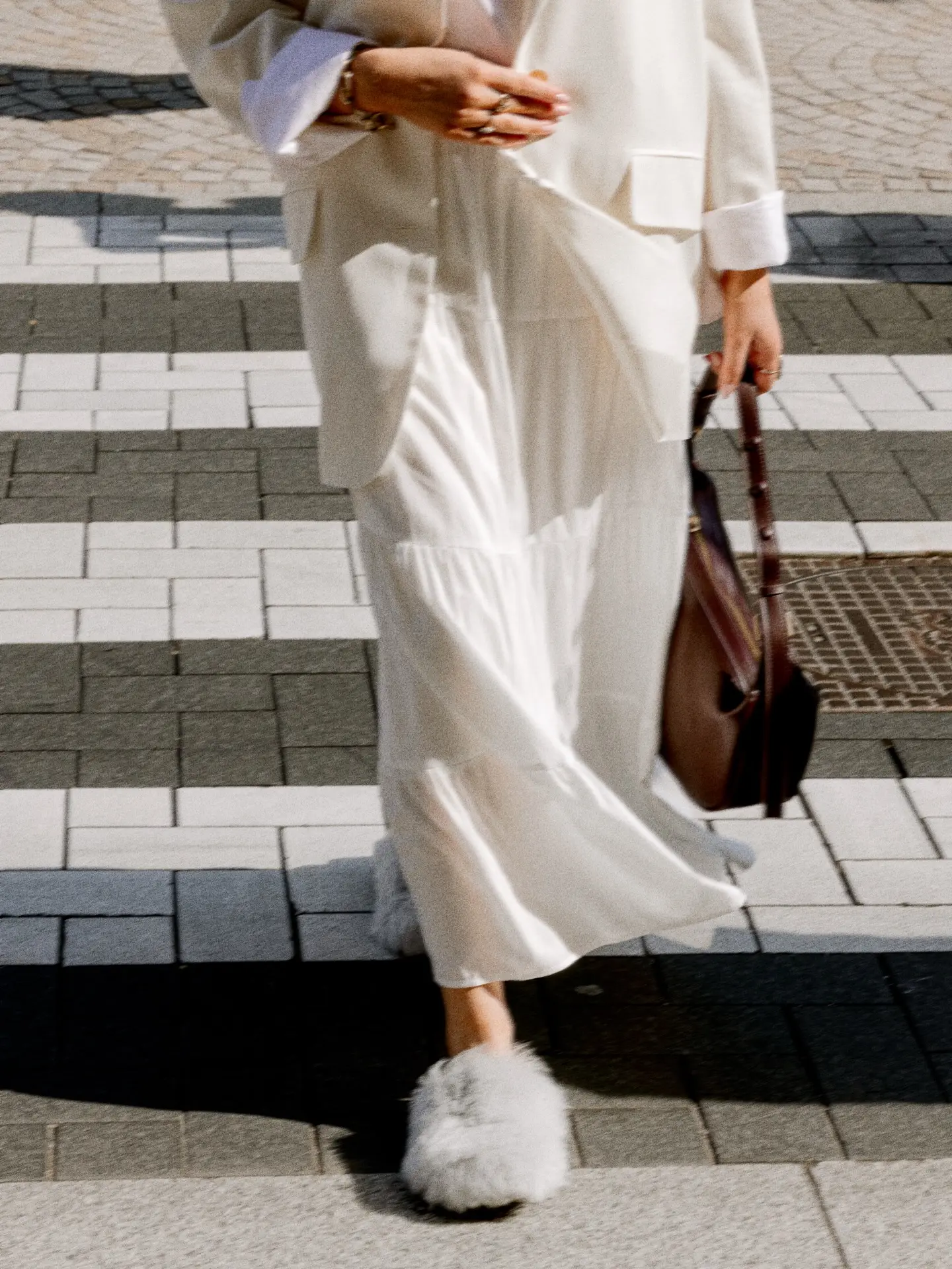 Model in witte blazer en rok met bruine tas en pluizige pantoffels op een gestraat plein