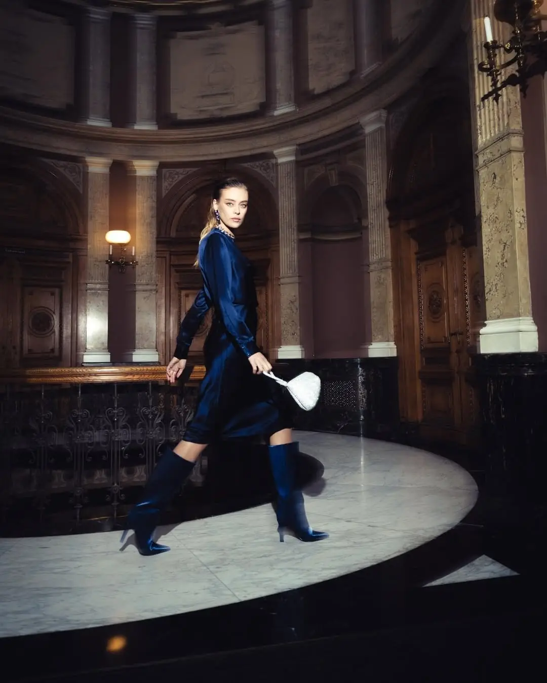 Model in donkerblauwe outfit en hoge laarzen loopt door een monumentale hal
