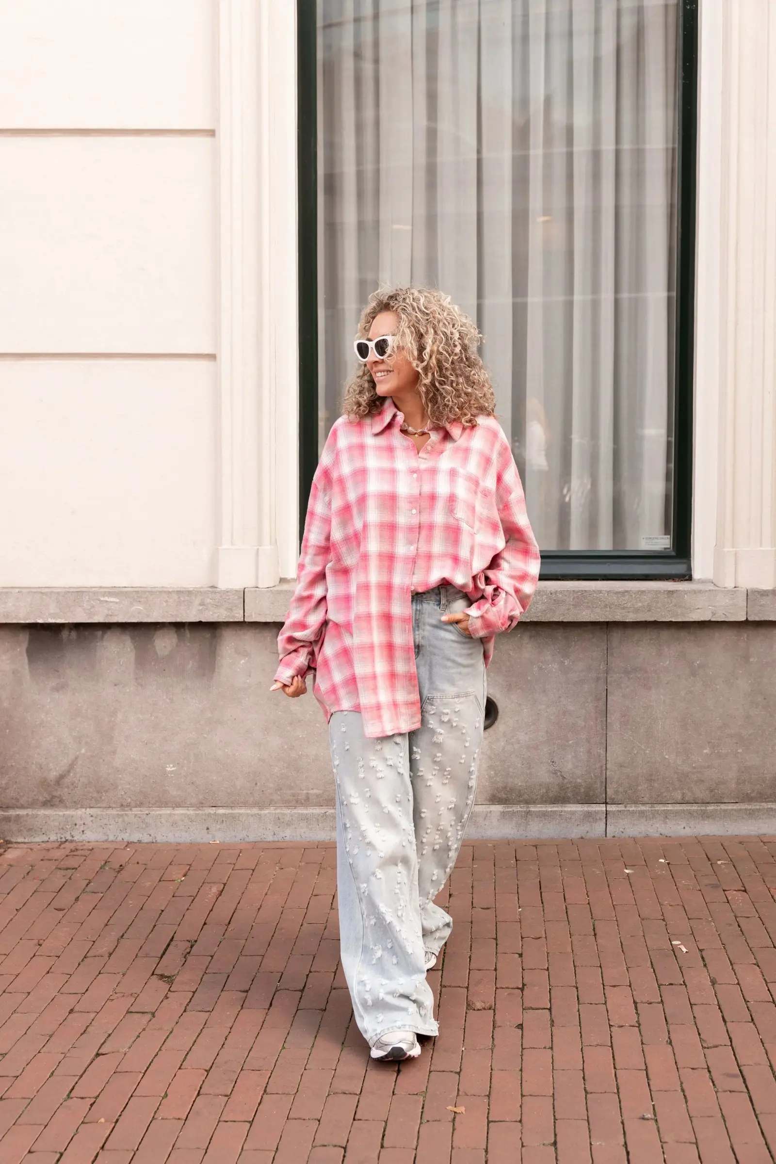 Vrouw met blond krullend haar en zonnebril in roze geruit overhemd en lichte jeans voor een gevel