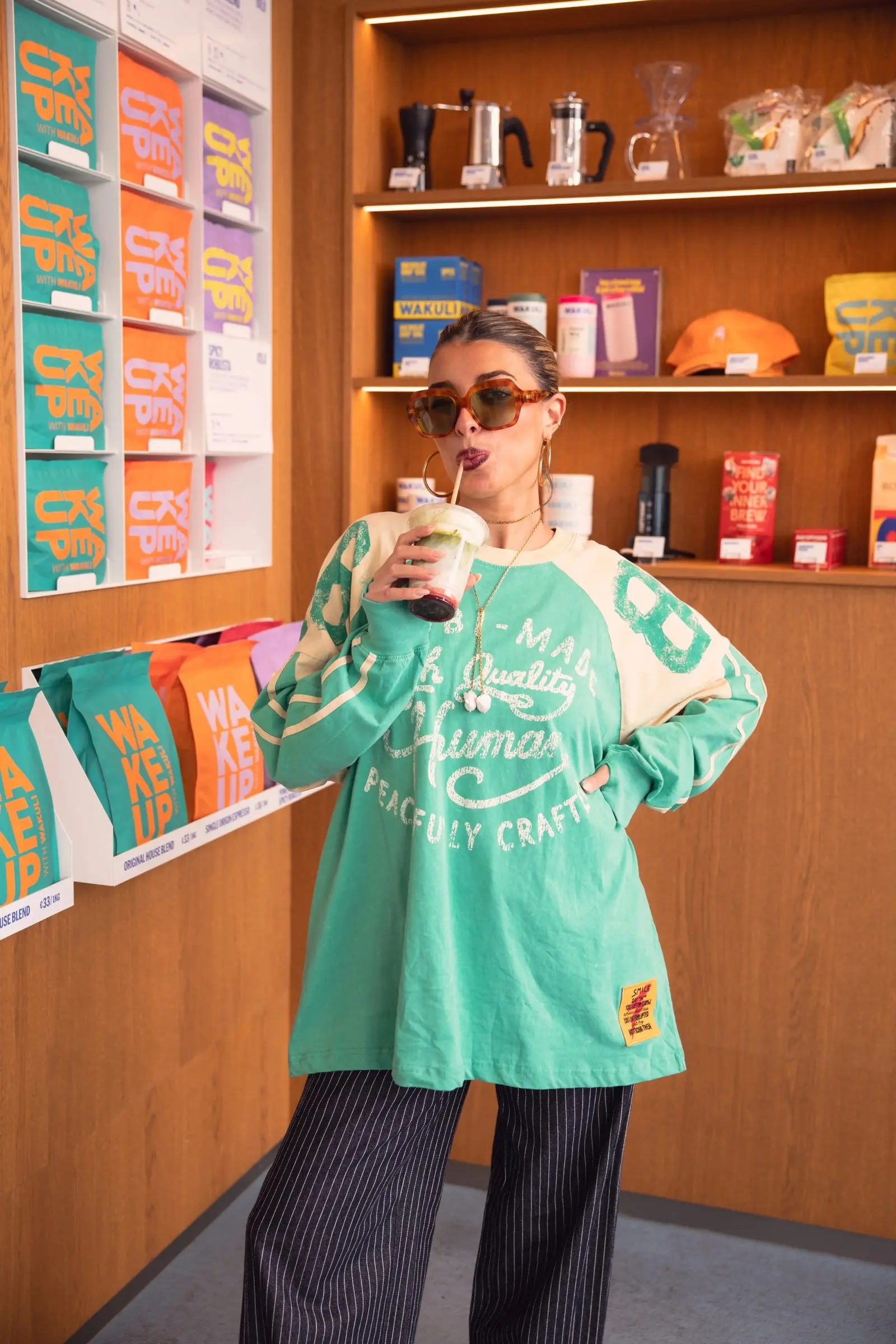 Vrouw met zonnebril en een drankje in een groen oversized shirt in een winkel met koffiezakken